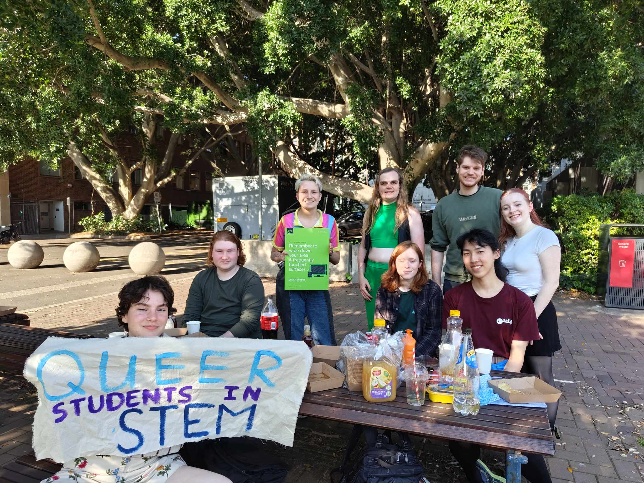 A group of people sit at a picnic table holding a sign that reads Queer Students in STEM