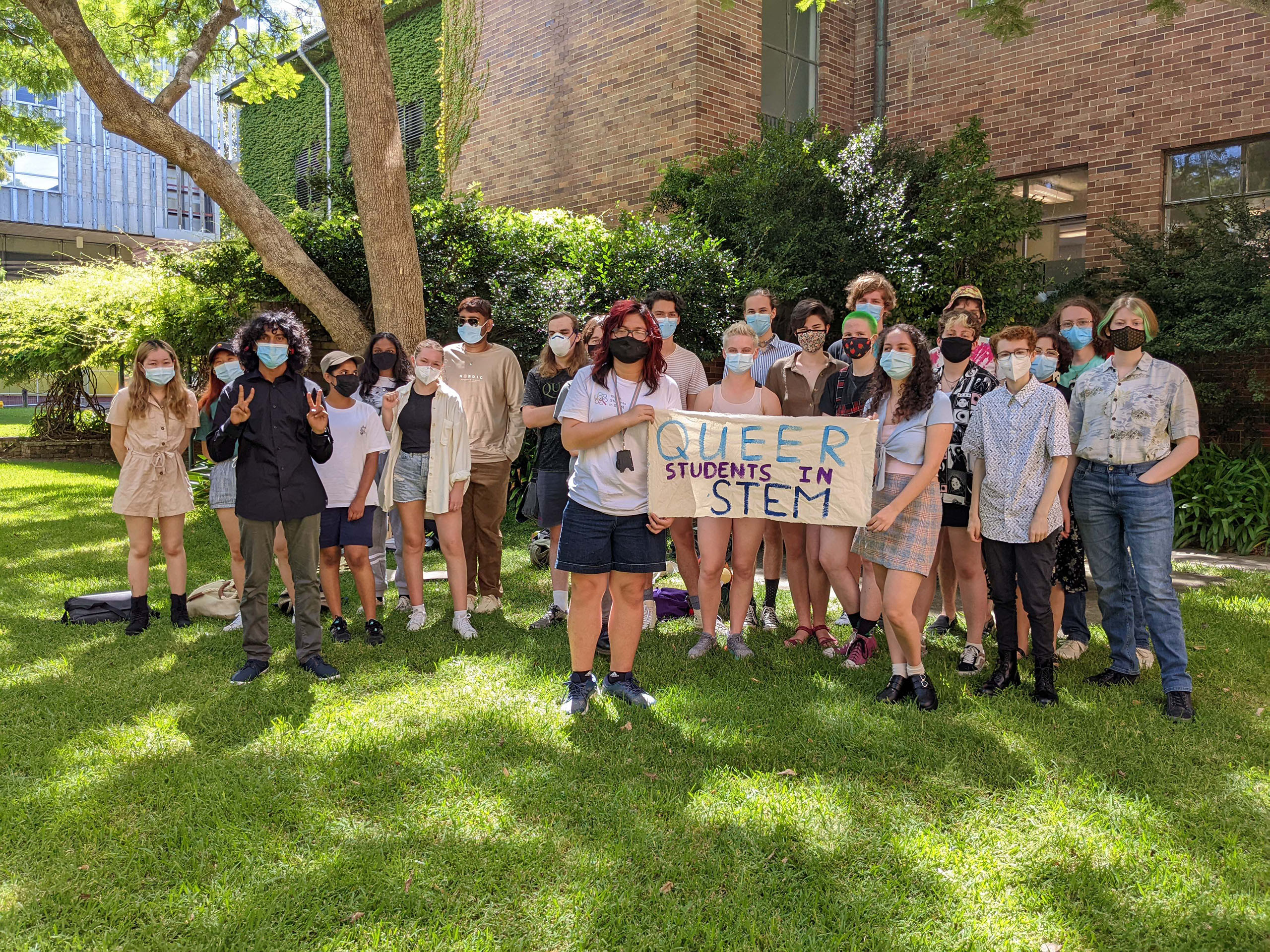 A group of people wearing masks on a lawn holding a sign that reads Queer Students in STEM