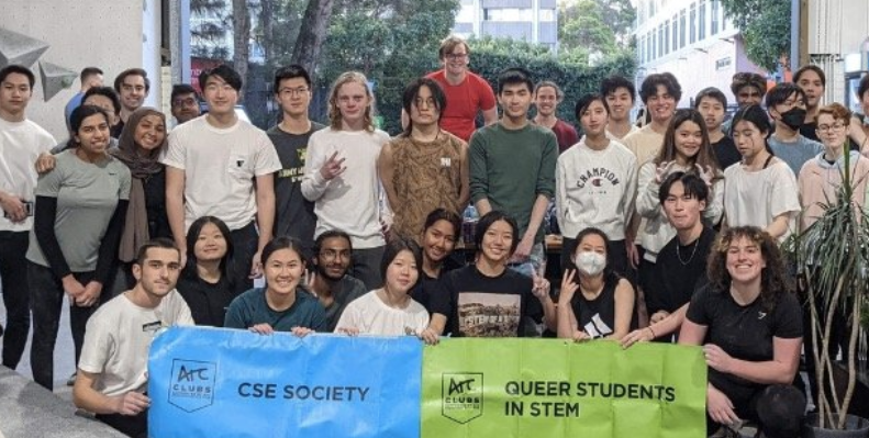 People standing in a climbing gym with two signs reading CSE society and Queer Students in STEM